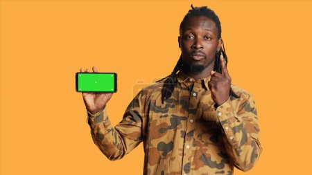 Photo for Cheerful adult holds smartphone with greenscreen in studio, presenting isolated copyspace display on mobile phone layout. Young person showing blank chromakey mockup on device. - Royalty Free Image