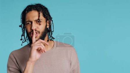 Photo for Annoyed Middle Eastern man doing shushing hand gesturing, irritated by noise, having negative mood. Assertive person placing finger on lips, doing quiet sign gesture, studio background, camera B - Royalty Free Image