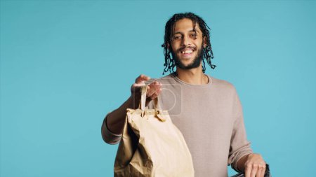 Téléchargez les photos : Portrait d'un livreur de pizza prenant un sac à lunch en papier à emporter dans un sac thermique. messager moyen-oriental sortir le repas du sac à dos alimentaire, isolé sur fond de studio bleu, caméra A - en image libre de droit