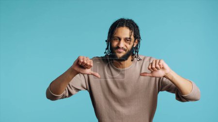 Upset Middle Eastern man showing thumbs down sign gesturing, disapproving, offering negative feedback. Disappointed BIPOC person doing rejection hand gesture, studio background, camera A