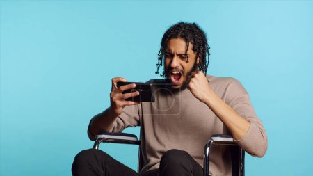 Joyful person in wheelchair playing videogames on smartphone, having positive burst of emotion after defeating opponents. Ecstatic gamer winning game on phone, studio background, camera A