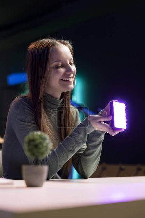 Happy young influencer in studio using camera to capture live broadcast, reviewing mini RGB lights. Joyful media star films electronics haul, presenting wireless LED lighting device