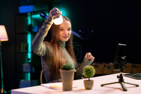 Upbeat influencer in neon lit studio filming review of light bulb able to change into different colors. Smiling gen Z tech expert presents smart home automation technology controlled by voice commands