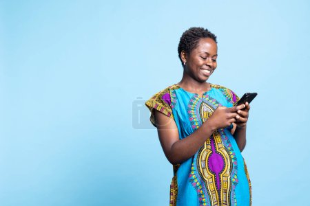 Téléchargez les photos : Une personne afro-américaine envoyant des SMS sur une application smartphone, naviguant sur des sites Web en ligne sur fond bleu. Jeune femme en costume ethnique traditionnel naviguant sur le réseau mobile en studio. - en image libre de droit