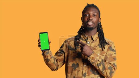 Photo for Smiling man points to smartphone with greenscreen on camera, showing blank mockup template on mobile device display. Young adult presenting isolated chromakey layout on phone screen. - Royalty Free Image