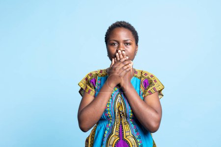 African american model covering her mouth to be silent, recreating three wise monkeys symbol to keep quiet in front of the camera. Woman showing inspirational traditional sign in studio.