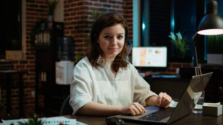 Photo for Portrait of smiling accountant researching key data for company project, doing tasks in office overnight. Happy employee sitting at desk in front of laptop, looking over accounting figures, camera A - Royalty Free Image