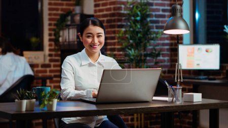 Photo for Portrait of smiling asian businesswoman doing computer tasks for team project in office. Cheerful employee working late at night, typing on laptop keyboard next to coworker, camera B - Royalty Free Image