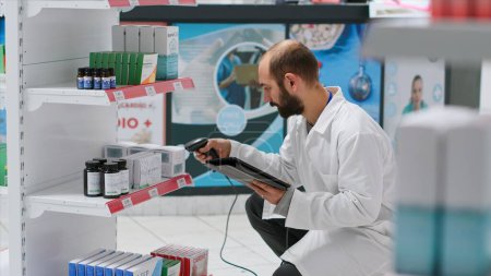 Photo for Pharmaceutical helper monitors the medication stock on shelves, scanning boxes that include prescribed medicine and cures at the clinic. Worker records medical supplies using tablet and scanner. - Royalty Free Image