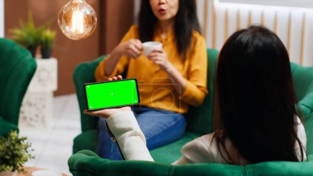 Asian traveler using smartphone with blank greenscreen at hotel, looking at template with chromakey and isolated mockup on display. Woman using phone and relaxing in lounge area.