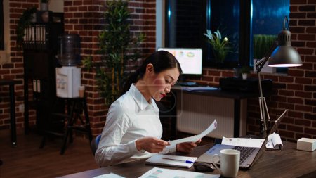 Asian employee reluctantly doing work in office late at night, bored of checking financial data charts on laptop. Businesswoman annoyed by having to check paperwork, feeling stressed and overworked