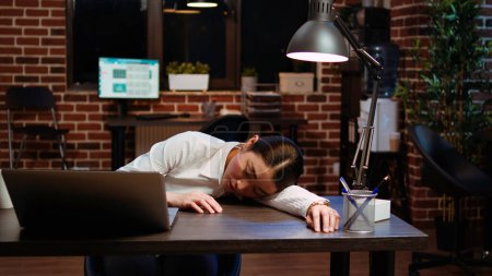 Tired employee sleeping on workspace desk after struggling all night to finish writing emails. Drowsy businesswoman napping in office after working overnight, asleep at laptop, camera A
