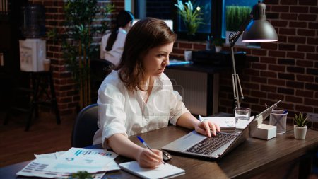 Worker writing financial business details on notepad, crosschecking with information on laptop. Businesswoman in brick wall office focused on finishing company project, camera A