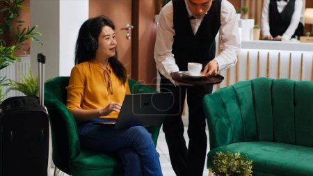 Photo for Asian woman receiving coffee while she works on laptop, sitting in lounge area at five star hotel and waiting for registration. Waiter serving drinks for guest relaxing, hospitality industry. - Royalty Free Image