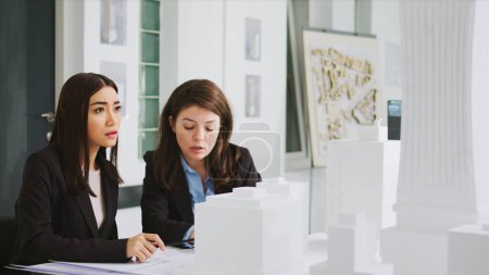 Architectural agency workers taking notes on blueprints, calculating real scale measurements to ensure the perfect elements for the construction team. Women developers studying 3d printed model.