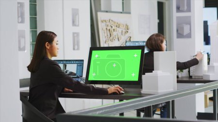 Téléchargez les photos : Architecte utilisant un ordinateur avec écran vert au poste de travail dans le bureau de l'agence, en regardant le modèle de maquette vierge sur l'écran. Ingénieur employé dans une petite entreprise immobilière. Trépied. - en image libre de droit