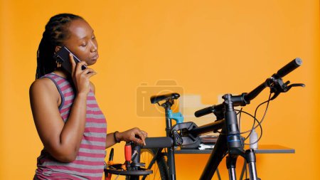 Mujer afroamericana hablando por teléfono con el mecánico, pidiendo ayuda en la reparación de la bicicleta dañada, fondo del estudio. Persona BIPOC en llamada telefónica con ingeniero de bicicletas, cámara B