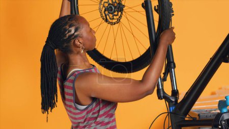 Téléchargez les photos : Femme faisant de l'entretien de vélo dans le studio atelier d'arrière-plan magasin, à la recherche de substitut à roue défectueuse. Le technicien fixe les pneus défectueux et entretient le dérailleur arrière endommagé - en image libre de droit