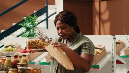 Customer pouring a type of pasta in ecological paper bag used to buy bulk products at zero waste store. Vegan woman searching for organic bio food alternatives at natural farming shop.