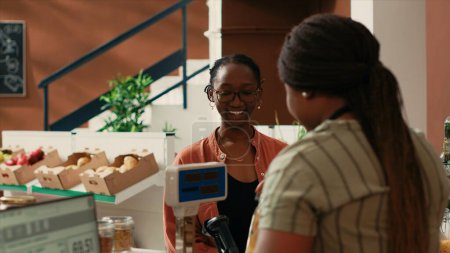 Fournisseur local pondérant les produits en vrac en vente pour l'acheteur féminin, propriétaire de petite entreprise à l'épicerie du quartier. Femme afro-américaine vendant pâtes et sauces dans des bocaux en verre réutilisables.