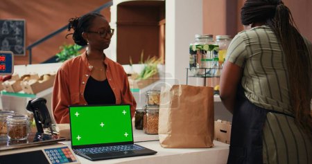 Photo for Retailer having laptop that shows greenscreen on display at cash register, giving eco products to client at market. Woman using computer with isolated chromakey template, mockup layout. Tripod shot. - Royalty Free Image