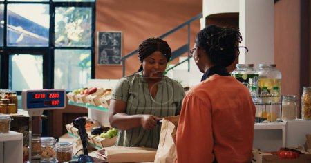 Photo for African american client paying for organic food using card, buying ethically sourced bio fruits and vegetables with pos payment at supermarket checkout. Woman purchasing produce. Handheld shot. - Royalty Free Image