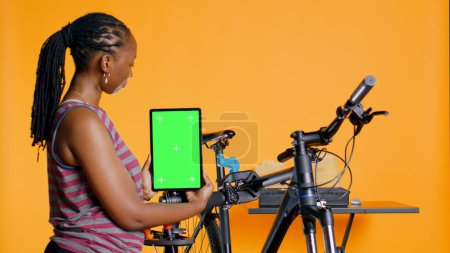 Foto de Mujer sostiene tableta de pantalla verde, utilizándolo para anunciar tienda de reparación de bicicletas, marcador de posición vacío para el logotipo de la empresa, fondo de estudio. Mecánico que promueve el servicio de fijación de bicicletas con dispositivo de maqueta, cámara B - Imagen libre de derechos