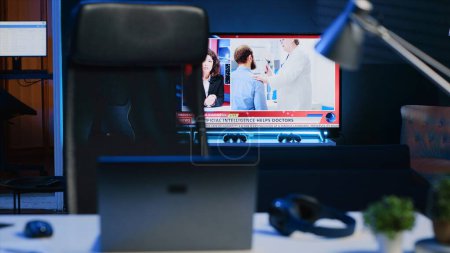 Photo for Smart TV left open on news broadcast in empty home office with desk filled with business items. Television set showing VOD channel with Laptop, headphones, plants in blurry foreground, camera A - Royalty Free Image