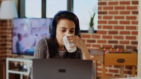 Photo for Indian woman drinking coffee in the morning and watching TV show at home using headphones. Matinal person waking up with hot beverage while enjoying television series on laptop, camera B - Royalty Free Image