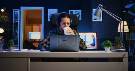 Foto de Hombre caucásico sentado en la silla en el escritorio de la oficina en el apartamento iluminado de neón, trabajando y escuchando música. Teletrabajador con auriculares en la sala de estar iluminada RGB, haciendo su trabajo de forma remota - Imagen libre de derechos