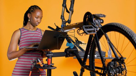 Téléchargez les photos : Ingénieur afro-américain effectuant l'entretien du vélo dans un atelier de réparation d'arrière-plan, en regardant sur la liste de maintenance d'ordinateur portable. Contrôle professionnel des pièces de bicyclette nécessitant une fixation, caméra B - en image libre de droit