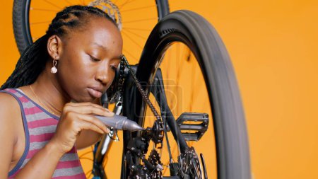BIPOC technician using specialized glue to repair broken bicycle chain, orange studio background. Professional applying adhesive on bike components during maintenance process, close up shot, camera B
