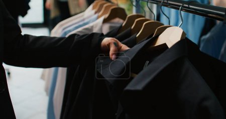 Photo for Classy woman examining all colorful shirts on display at clothing store, shopping for trendy brands to expand wardrobe. Customer looking at fashion items stored on hangers. Close up. Camera B. - Royalty Free Image