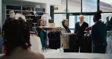 Photo for Store employee helping senior people with clothing suggestions, presenting new fashion collection items to couple in department store. African american employee makes recommendations. Camera B. - Royalty Free Image