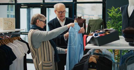 Photo for Senior couple shopping for formal outerwear at fashion boutique, looking for trendy elegant clothing items to create new outfits for elderly man. People searching for stylish merchandise. Camera B. - Royalty Free Image