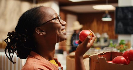 Comprador afroamericano disfrutando de aroma fresco de tomates en la tienda, oliendo productos recién cosechados colocados en cajas en el supermercado del vecindario. Mujer apoyando la agricultura local. Cámara 2.