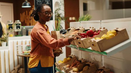 Comprador afroamericano que elige productos orgánicos de cajas, poniendo frutas y verduras en una bolsa de papel para comprar. Mujer comprando productos ecológicos naturales en el mercado de agricultores locales. Cámara 1.