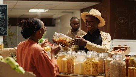 Farmer with apron recommending new homemade sauce for woman, showing organic pasta types and other pantry supplies. African american vendor presenting products to regular customer. Camera 1.