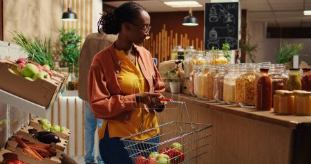 Cliente regular en busca de alternativas de alimentos ecológicos, mirando artículos a granel hechos en casa de origen ético en el supermercado local. Mujer vegana afroamericana yendo de compras. Cámara 2.
