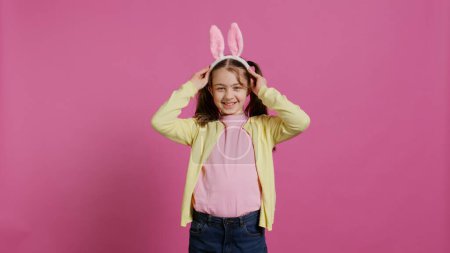 Téléchargez les photos : Adorable enfant mignon mettant des oreilles de lapin et agitant à la caméra, profitant Pâques dimanche célébration sur fond rose. Écolière souriante joyeuse avec des nattes disant bonjour. Caméra B. - en image libre de droit