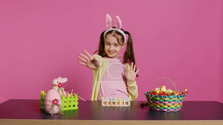Photo for Joyful schoolgirl with bunny ears waving hello in front of camera, sitting at a table with festive decorations and arrangements for easter sunday celebration. Young excited child. Camera B. - Royalty Free Image