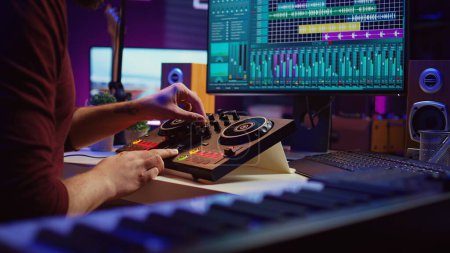 Artist editing sounds on mixing console in home studio, operating professional audio equipment to adjust volume levels on track recordings. Musician works with daw software an pre amp knobs, Camera B.
