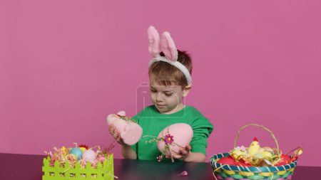 Kleine Kinder freuten sich über versteckte, handgefertigte Ornamente, die in Vorbereitung auf das Osterfest entstanden waren. Junge mit Hasenohren mit bemalten Dekorationen, einem Hasen und einem Ei. Kamera A.