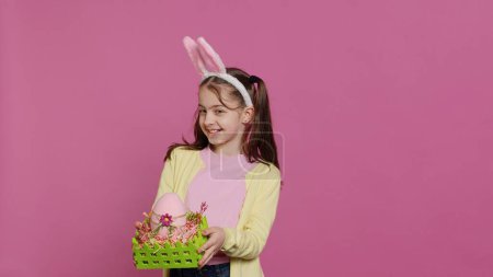 Photo for Smiling pleased girl presenting her handcrafted easter basket filled with painted eggs and other festive decorations for holiday celebration. Joyful small kid proud of her arrangement. Camera A. - Royalty Free Image