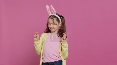 Photo for Lovely sweet girl posing in studio with fingers crossed and hoping to receive gifts for easter holiday celebration. Cheerful young child with bunny ears asking for good luck and fortune. Camera A. - Royalty Free Image