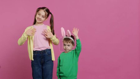 Photo for Joyful little kids waving in front of camera during easter holiday, smiling and wearing bunny ears. Brother and sister toddlers greeting someone in studio, adorable happy children. Camera A. - Royalty Free Image