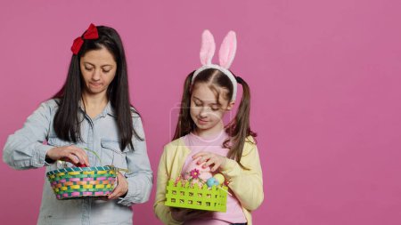 Photo for Joyful confident child and mother showing easter baskets on camera, decorating festive arrangements for spring holiday. Happy schoolgirl with bunny ears posing with her mom in studio. Camera A. - Royalty Free Image