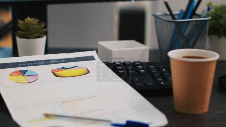 Photo for Financial report on desk next to paper cup of coffee in empty office, close up shot. Company files containing economic graphs and pie charts near hot beverage on table in workspace - Royalty Free Image