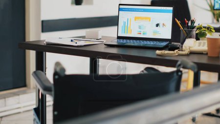 Wheelchair in office and laptop on desk showing business analytics. Medical chair in workplace offering accessibility to workers with disability and notebook with financial charts, panning shot