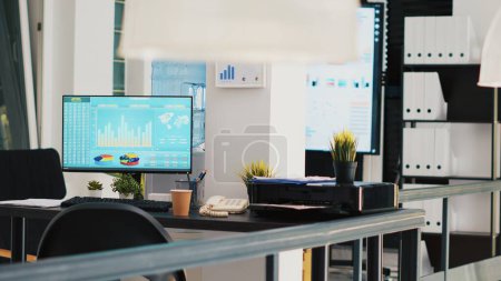 Büro mit Computermonitor auf dem Schreibtisch mit Börsennotierungen und Anzeige im Hintergrund mit Geschäftsdiagrammen. PC im Arbeitsbereich mit Handelsplattform-Indizes und Unternehmensdaten-Diagrammen im Hintergrund